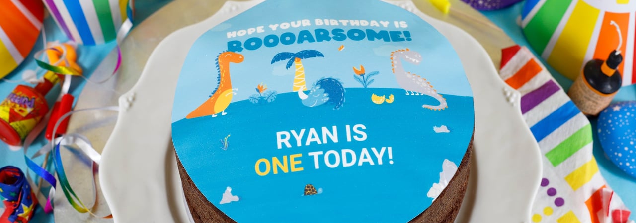 Boy's Birthday Cakes - Chocolate photo cake with blue dinosaur topper that says Ryan is one today - Blog Banner