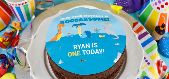 Boy's Birthday Cakes - Chocolate photo cake with blue dinosaur topper that says Ryan is one today - Blog Banner